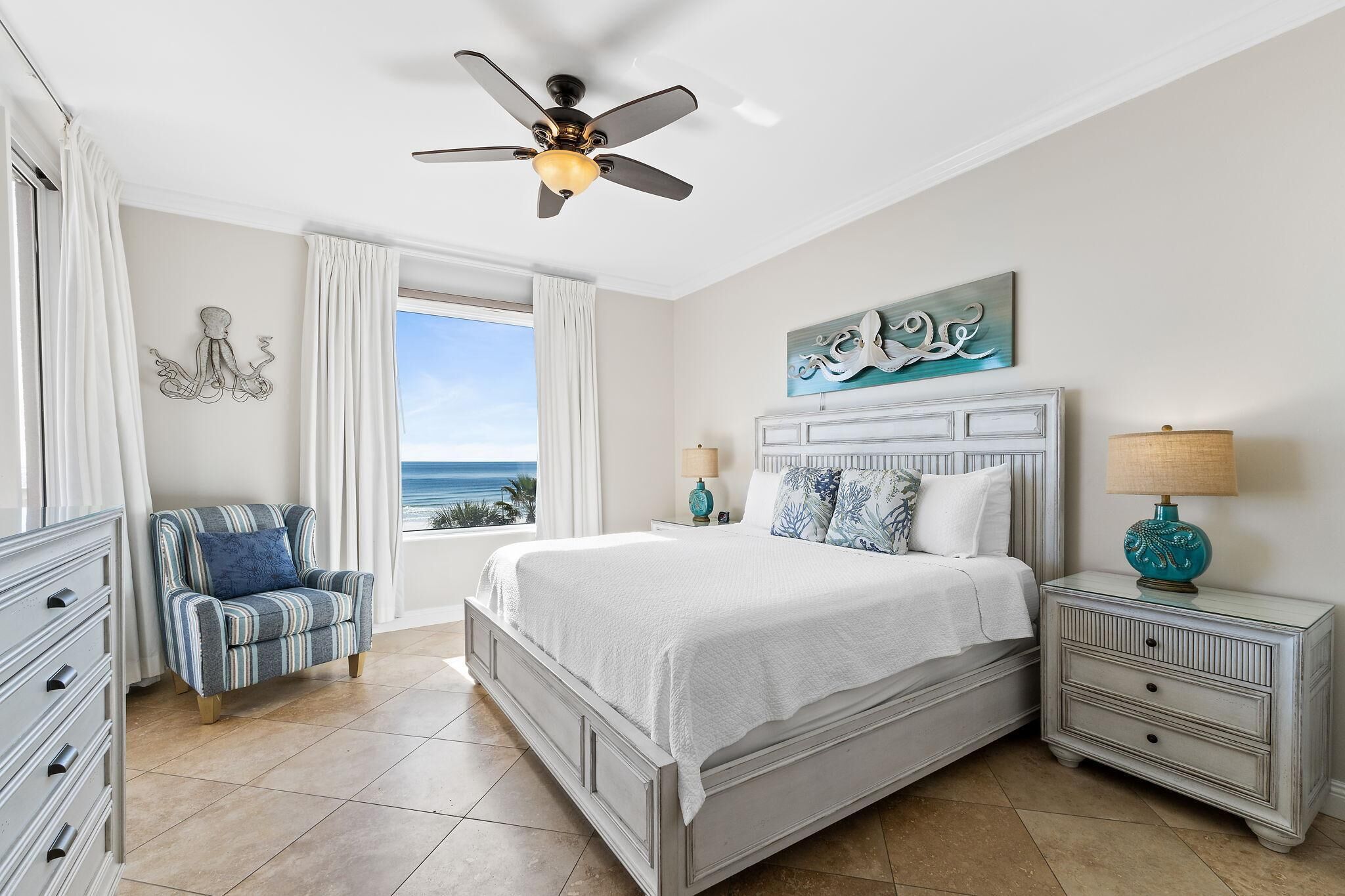 A bedroom with a bed, nightstands, a chair, a ceiling fan, and a view of the ocean.