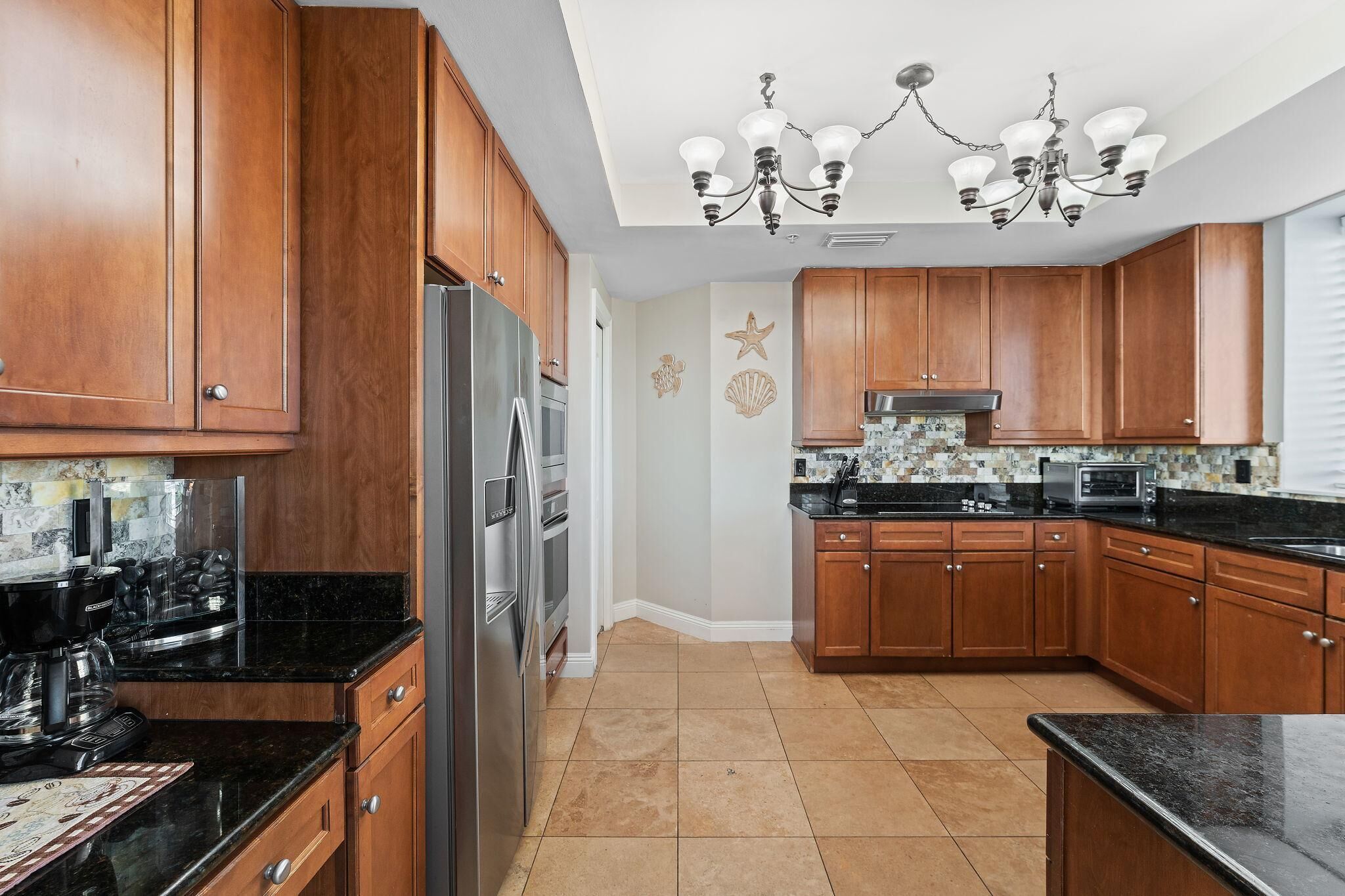 A modern kitchen with wooden cabinets, a stainless steel refrigerator, a black countertop, and a chandelier.