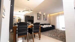 A modern living room with a black sofa, a dining table with chairs, and a large window with curtains. - PRIME LOK- SP FLAT VILA OLIMPIA (São Paulo)