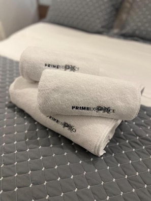 A bed with a quilted gray bedspread and two rolled white towels with 'PRIMEXPERIENCE' printed on them.