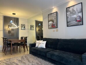 A modern living room with a dark grey sofa, a wooden dining table with chairs, and a chandelier. There are framed pictures on the walls, including one of a red double-decker bus.