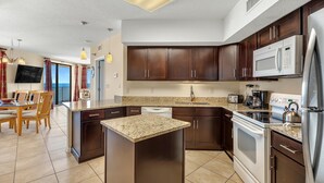 A modern kitchen with dark wood cabinets, a granite countertop, stainless steel appliances, and a dining area with a view. - Bluewater 1101 (Orange Beach)