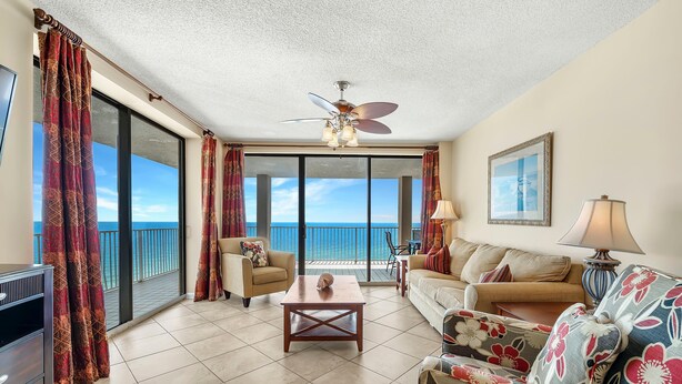 A spacious living room with a large window overlooking the ocean, a ceiling fan, and a sofa with floral upholstery. - Bluewater 1101 (Orange Beach)