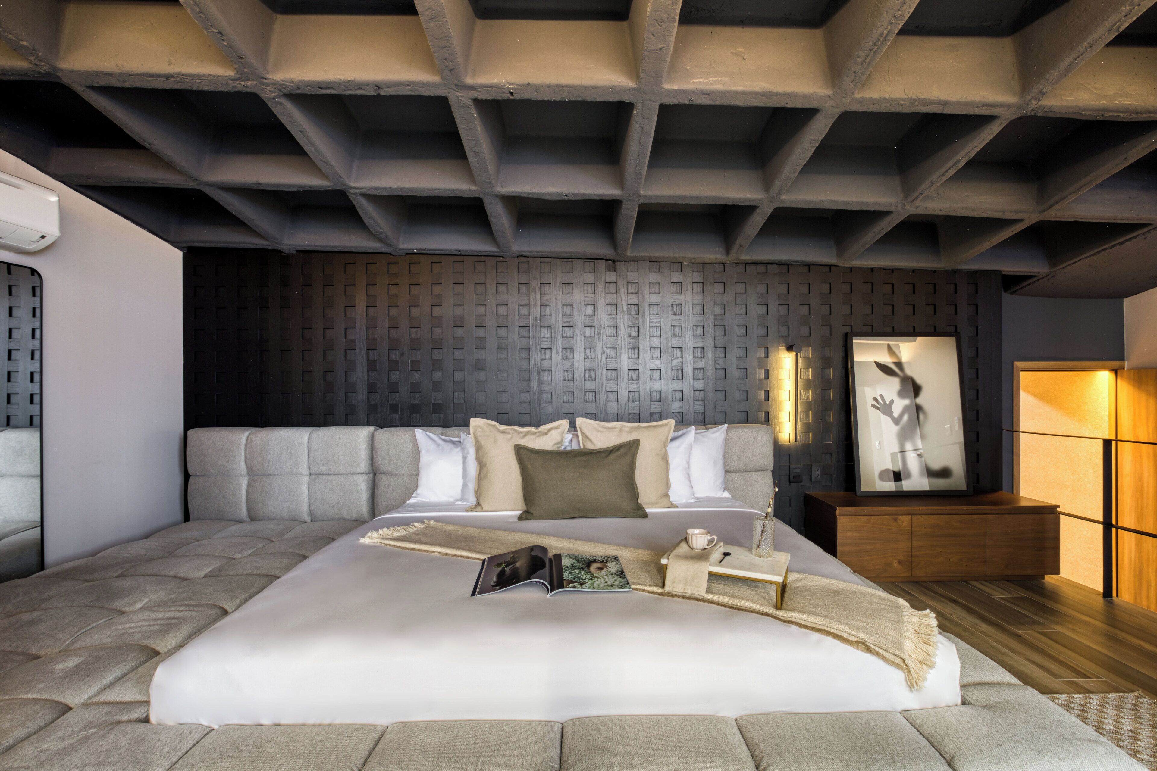 A modern bedroom with a large bed, a headboard, a bedside table, a nightstand, a wall-mounted light, and a framed picture.