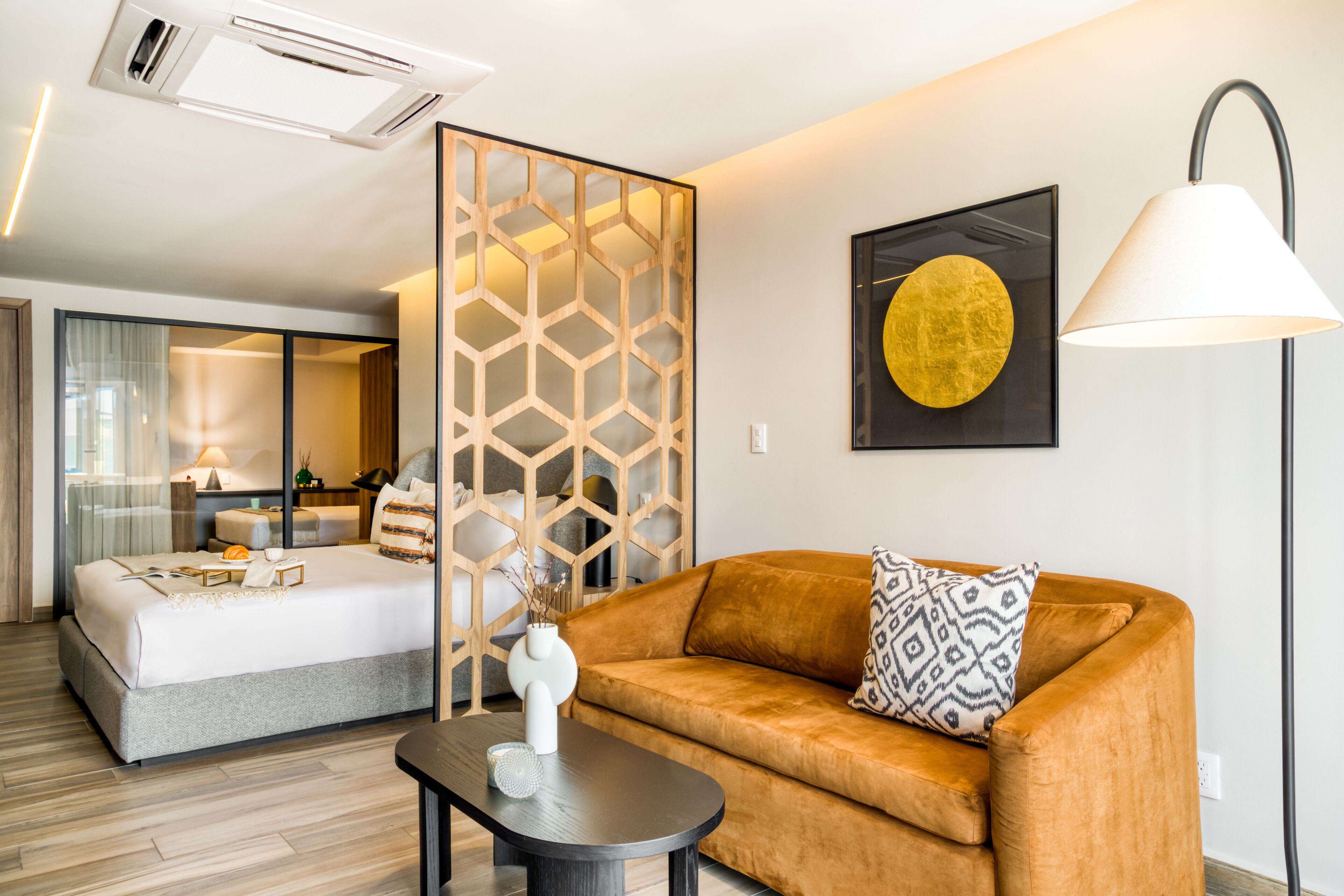 A modern hotel room with a bed, a sofa, a decorative partition, a lamp, and a framed artwork on the wall.