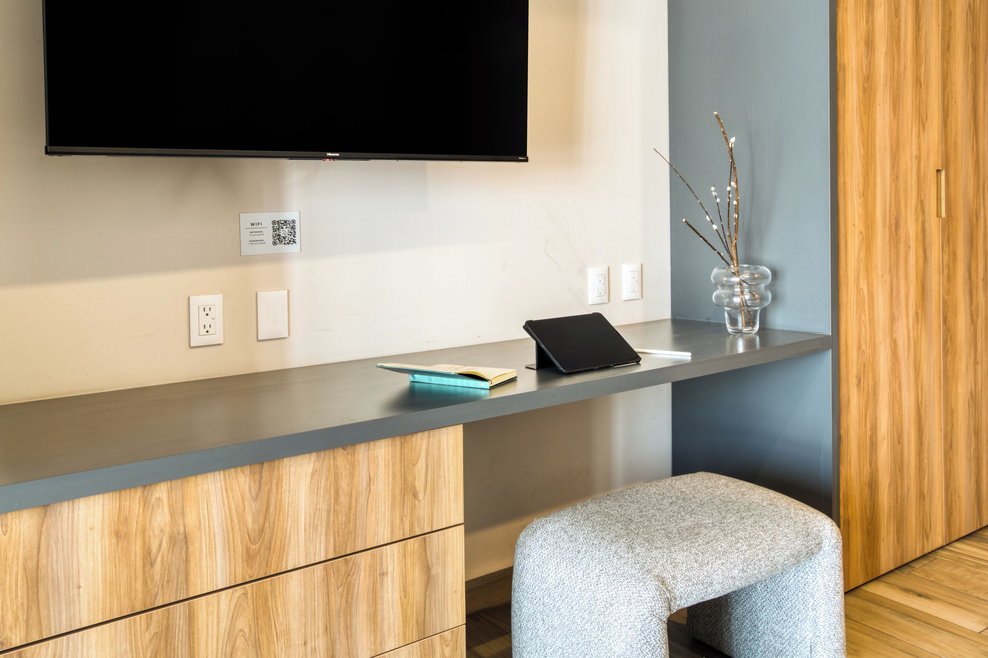 Una habitación moderna con televisión de pantalla plana en la pared, un escritorio de madera con una computadora portátil, un pequeño jarrón con ramas y un taburete acolchado gris.