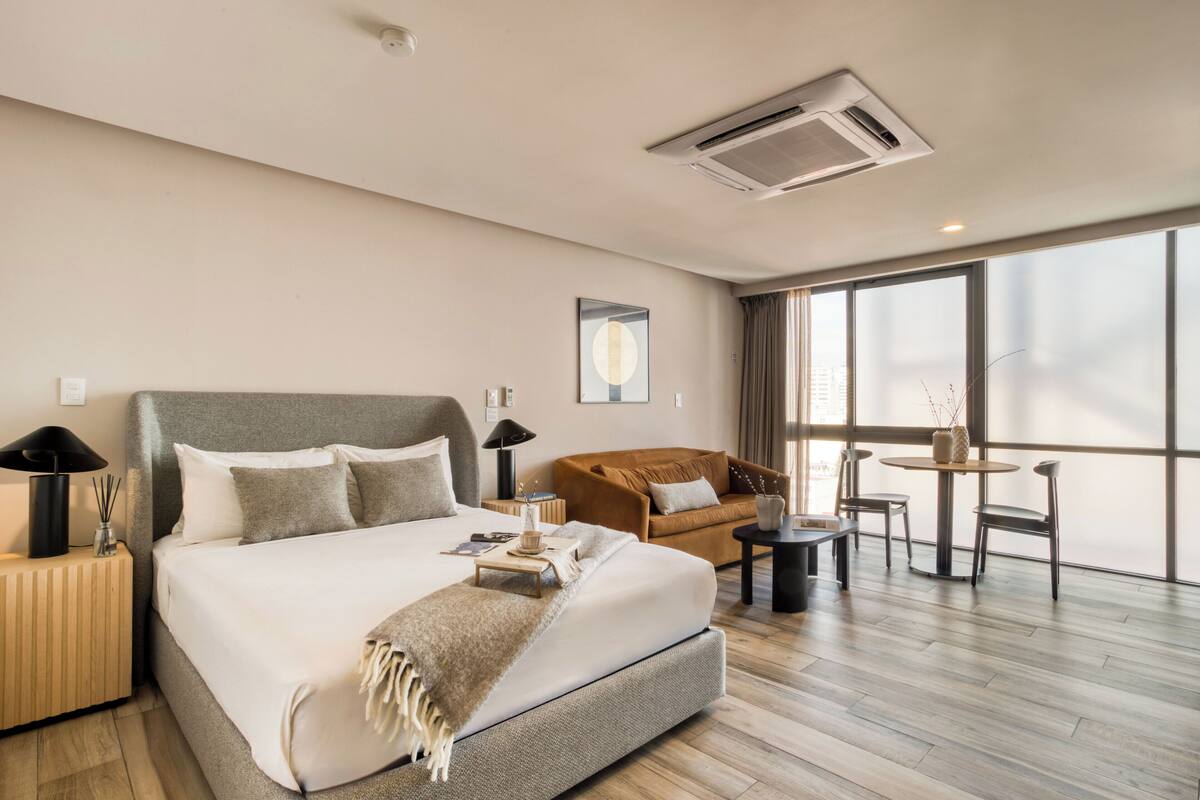 A modern hotel room with a large bed, a sofa, a small table, and a view of the city through large windows.