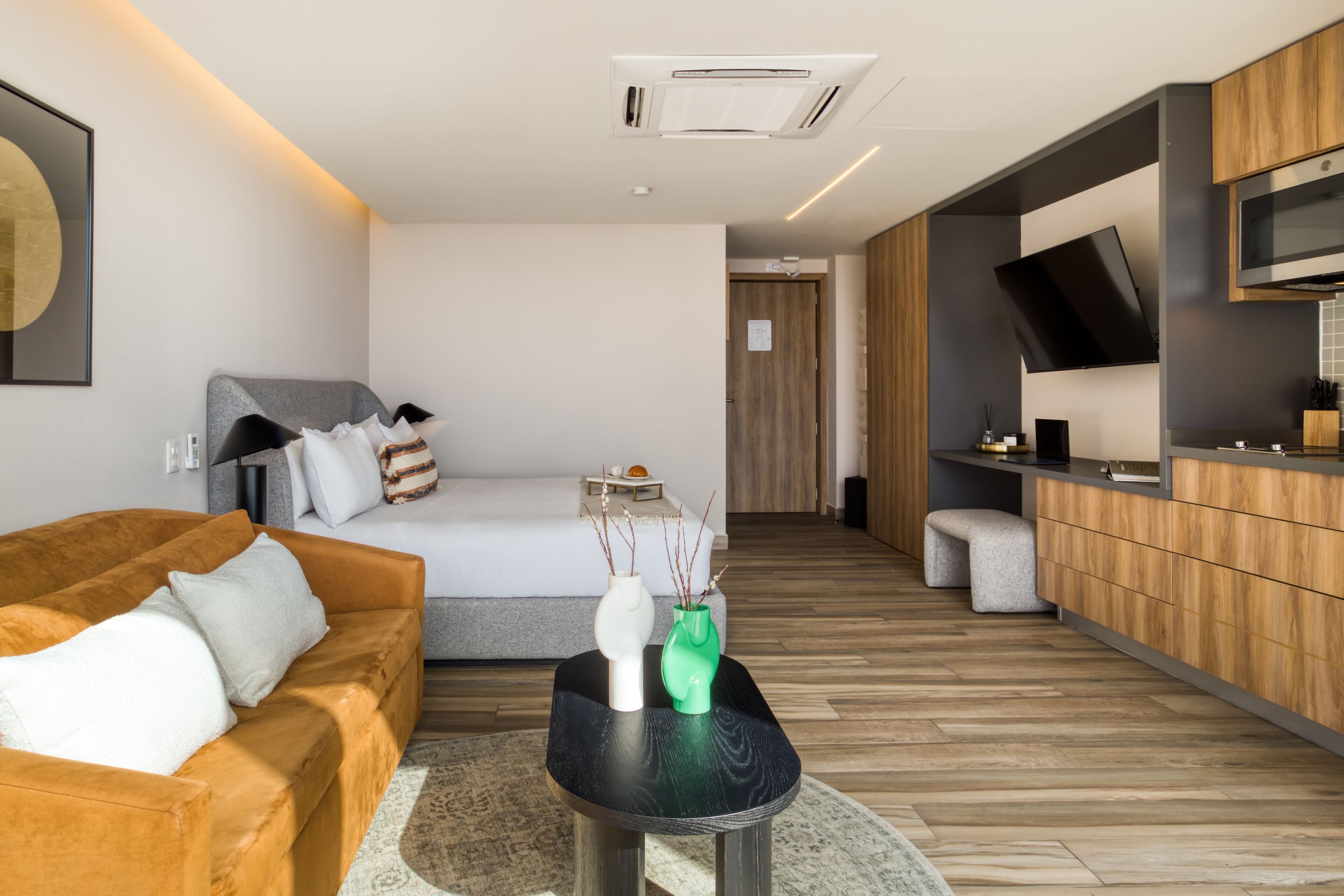 Een moderne hotelkamer met een bank, een bed, een kleine tafel met vazen, een keukenhoek met een televisie en een houten vloer.