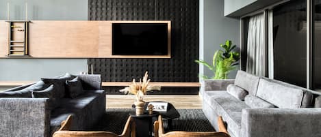 Uma sala de estar moderna com uma grande televisão de tela plana presa a uma parede texturizada, sofás cinzas, uma mesa de centro com objetos decorativos e uma planta.