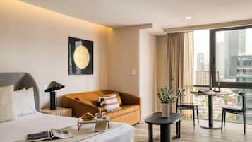 Habitación de hotel con cama, sofá, una mesita y vistas a la ciudad a través de amplias ventanas.