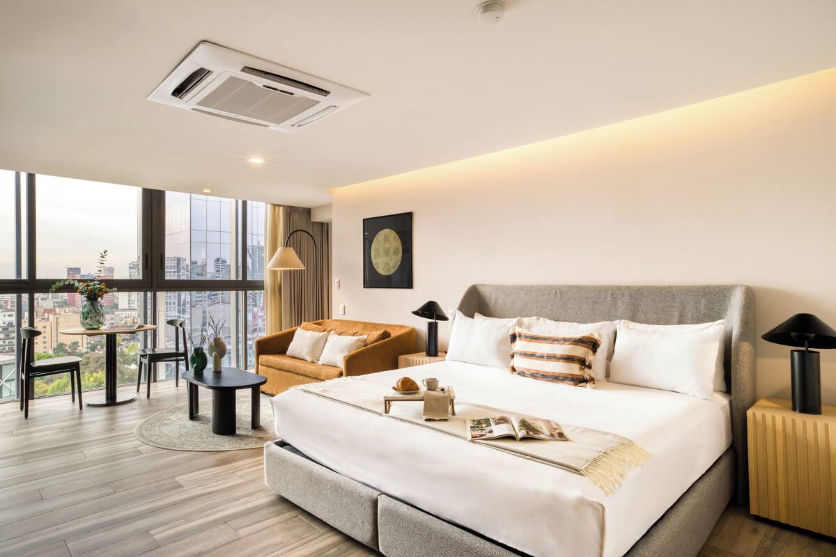 A modern hotel room with a large bed, a round dining table, a sofa, and a view of the city.