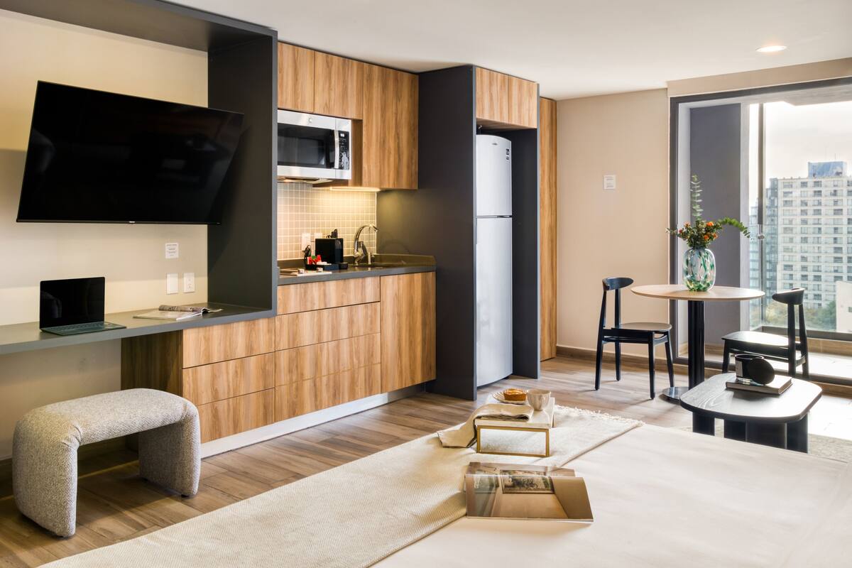 A modern kitchen with wooden cabinets, a microwave, and a refrigerator. There is a dining area with a table and chairs, and a large window offering a view of the city.