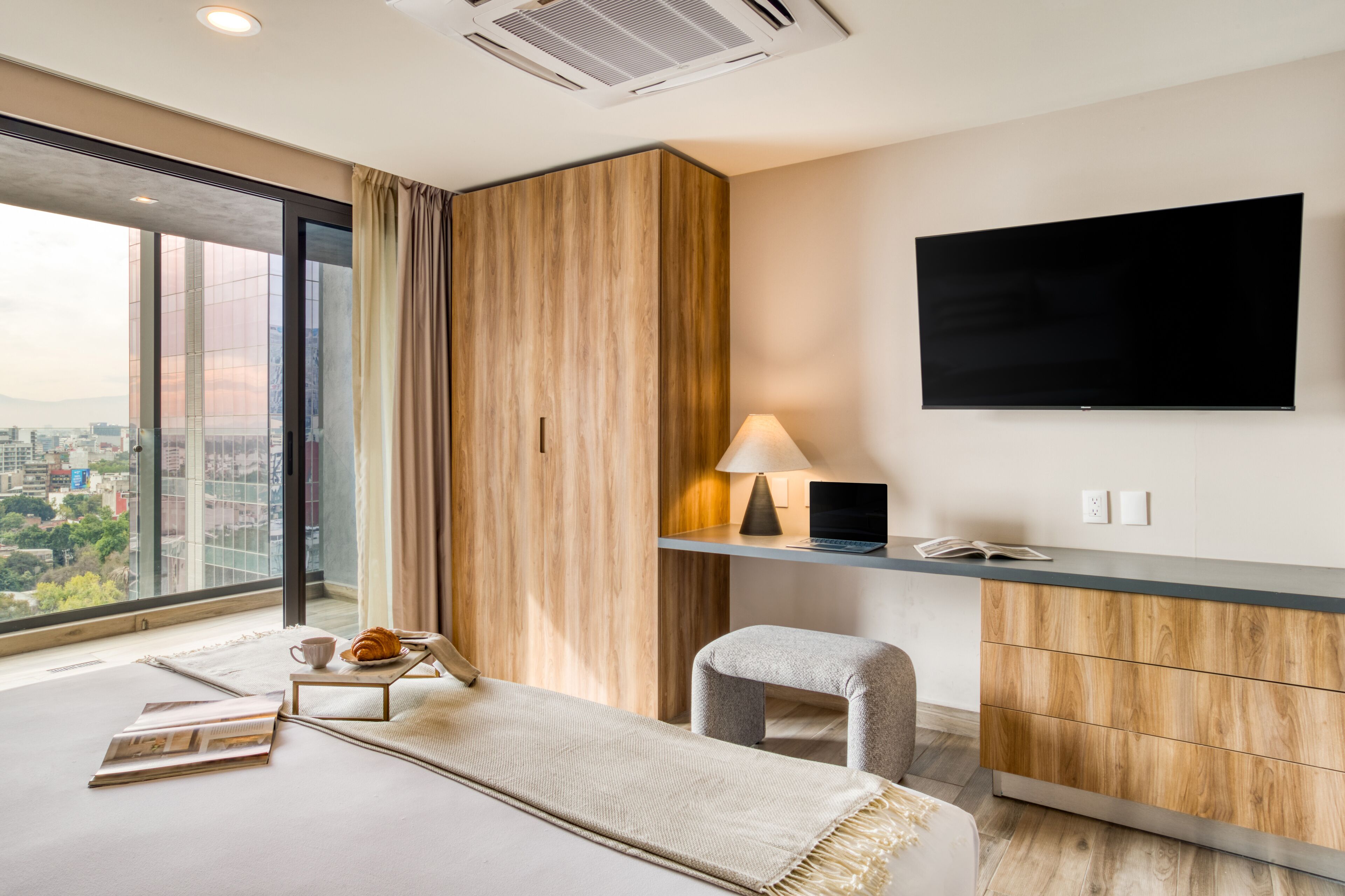 A modern hotel room with a large window, a flat-screen TV, a desk with a lamp, a sofa, and a small table with a croissant.