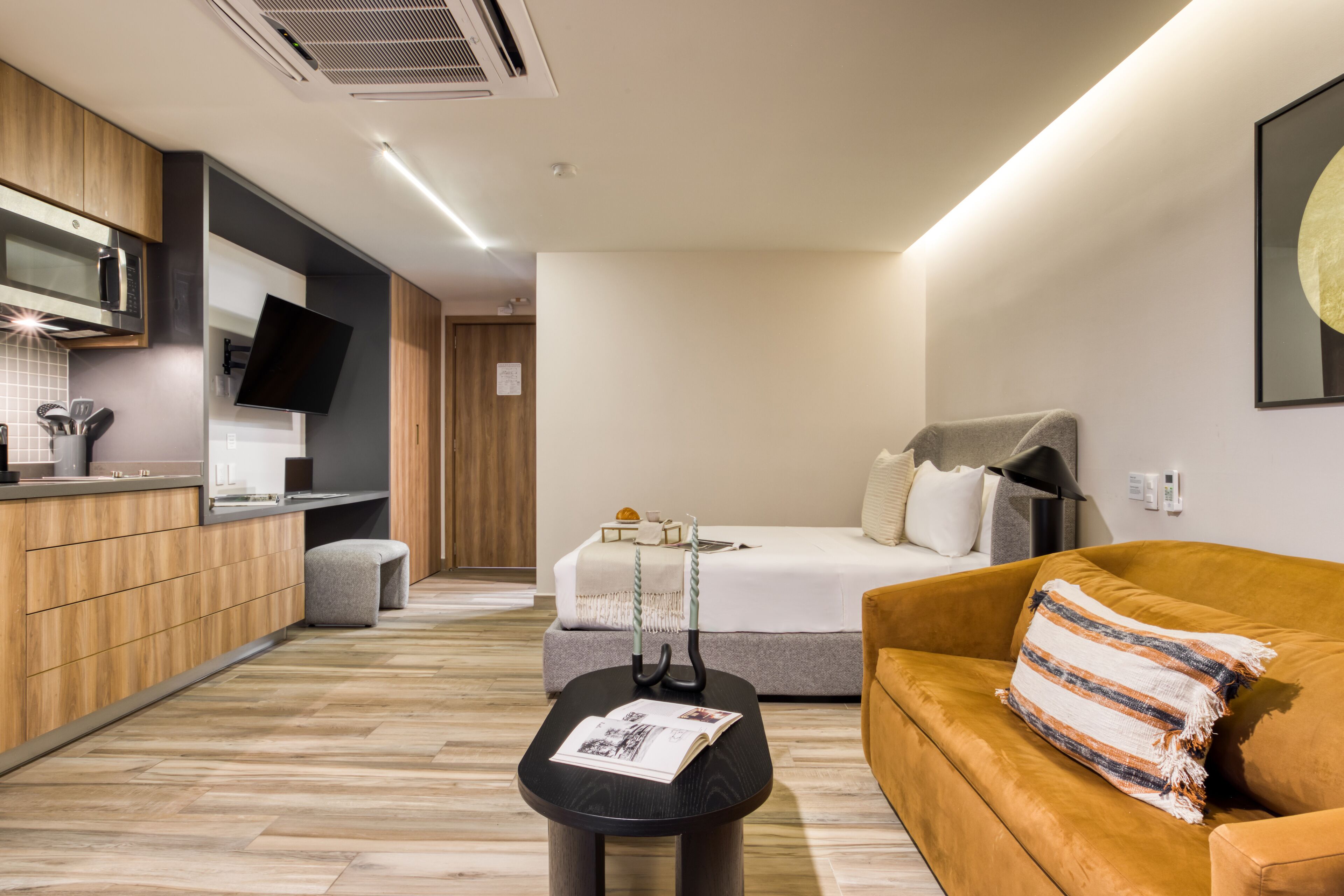 Una habitación de hotel moderna con cama, sofá, una pequeña mesa con una revista, televisor montado en la pared y una zona de cocina con armarios de madera y microondas.