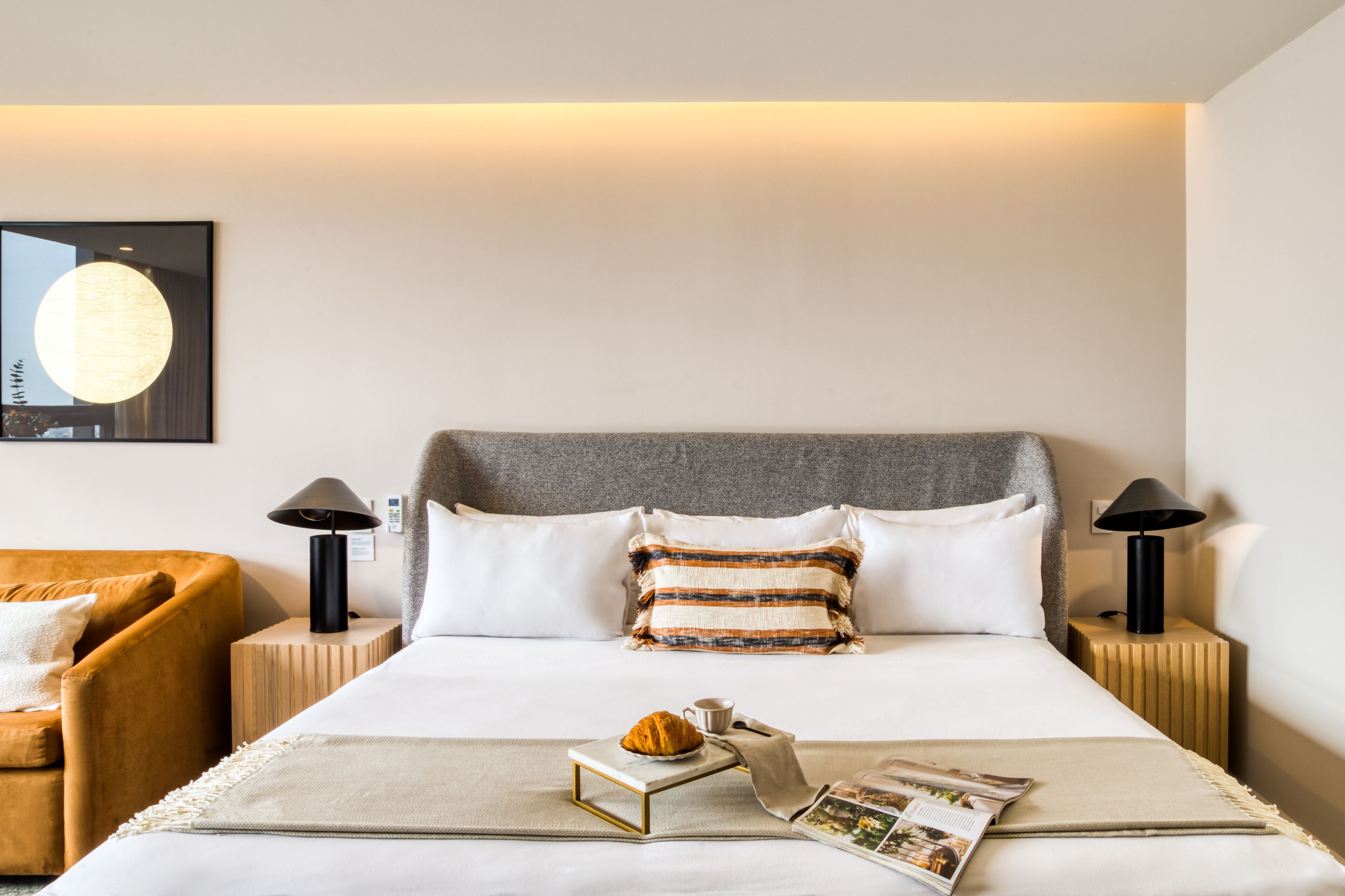 Habitación de hotel ordenada con una cama, una mesita auxiliar, una lámpara, una revista y un croissant.