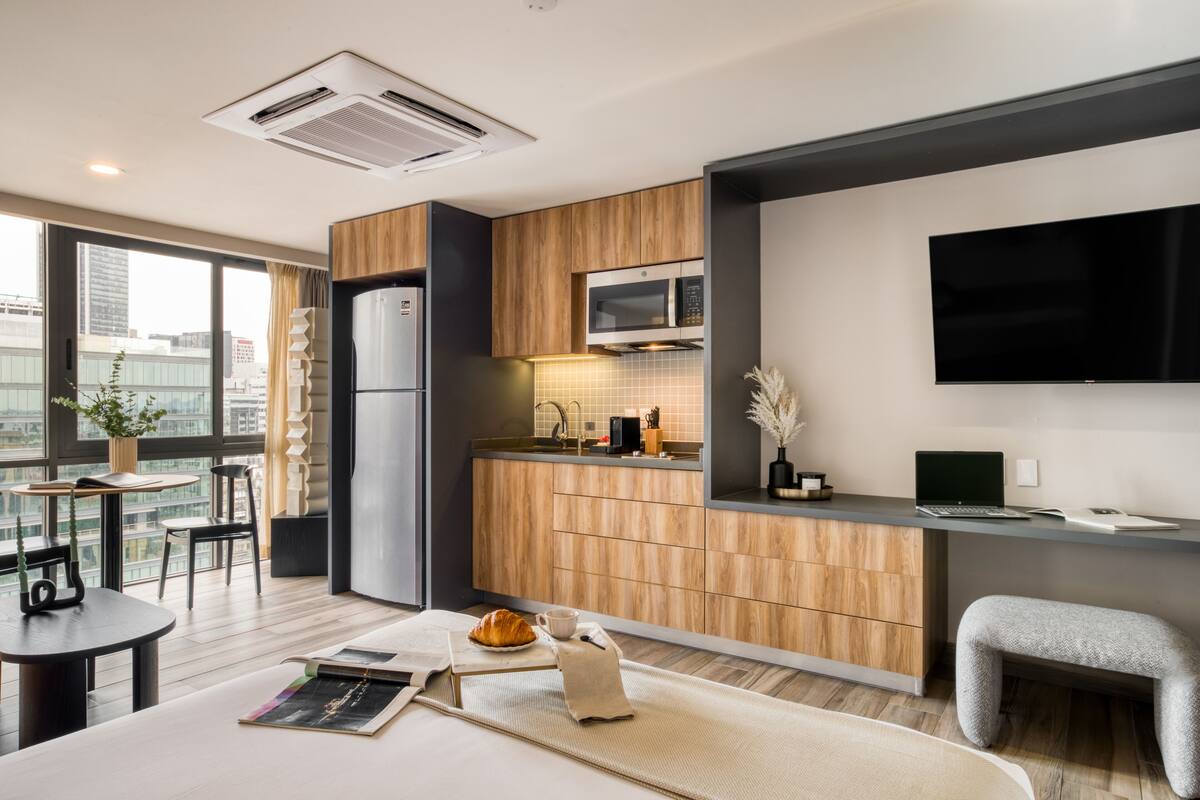 A modern kitchen with a microwave, refrigerator, and a flat-screen TV. There is a dining area with a table and chairs, and a small seating area with a bench.