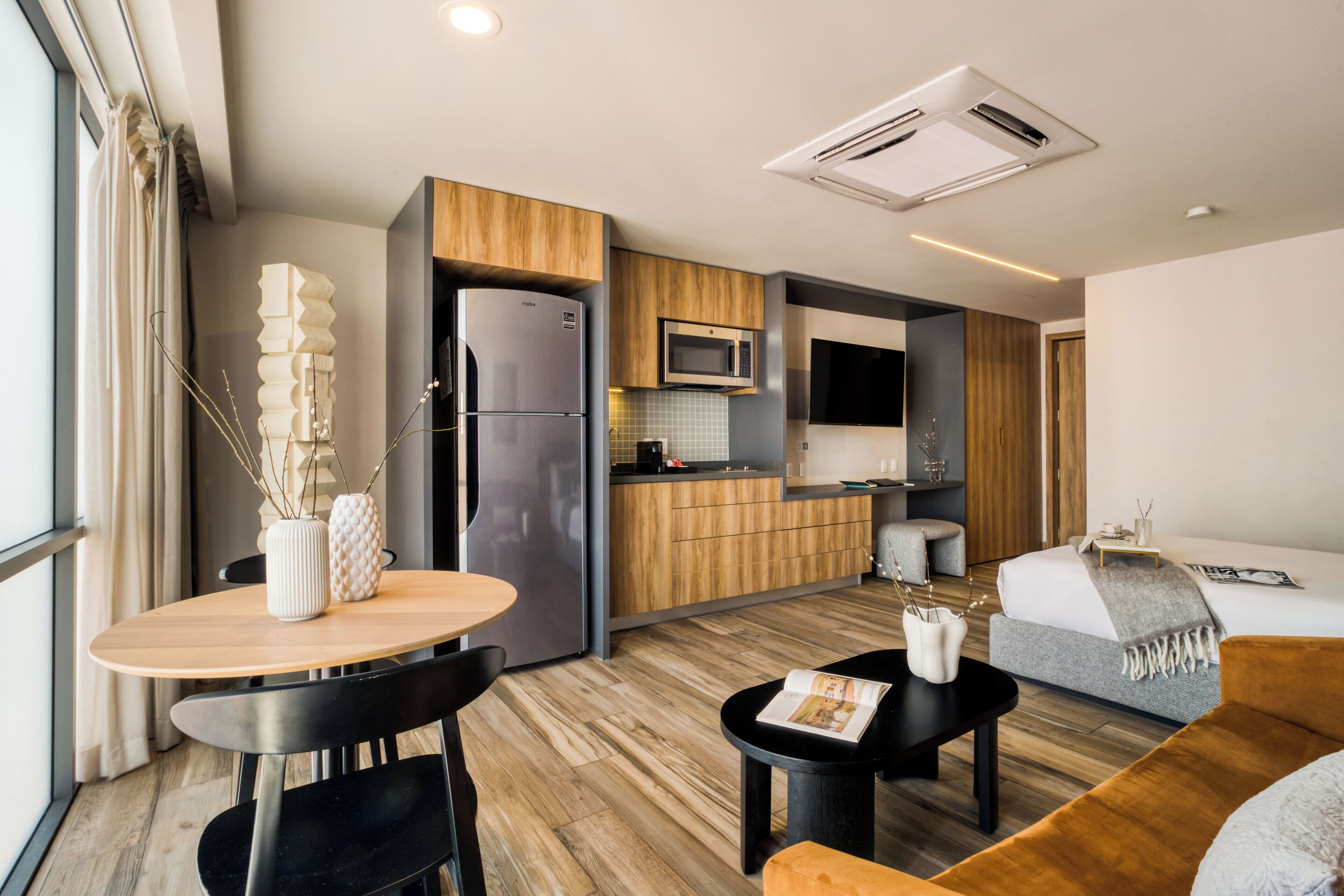 A modern kitchen with a refrigerator, microwave, and wooden cabinets. There is a dining area with a table and chairs, and a living area with a sofa and a coffee table.
