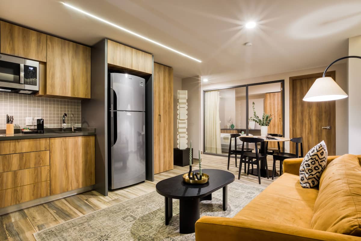 A modern kitchen with wooden cabinets, a built-in microwave, and a stainless steel refrigerator. There is a dining area with a table and chairs, and a sofa with a patterned cushion.