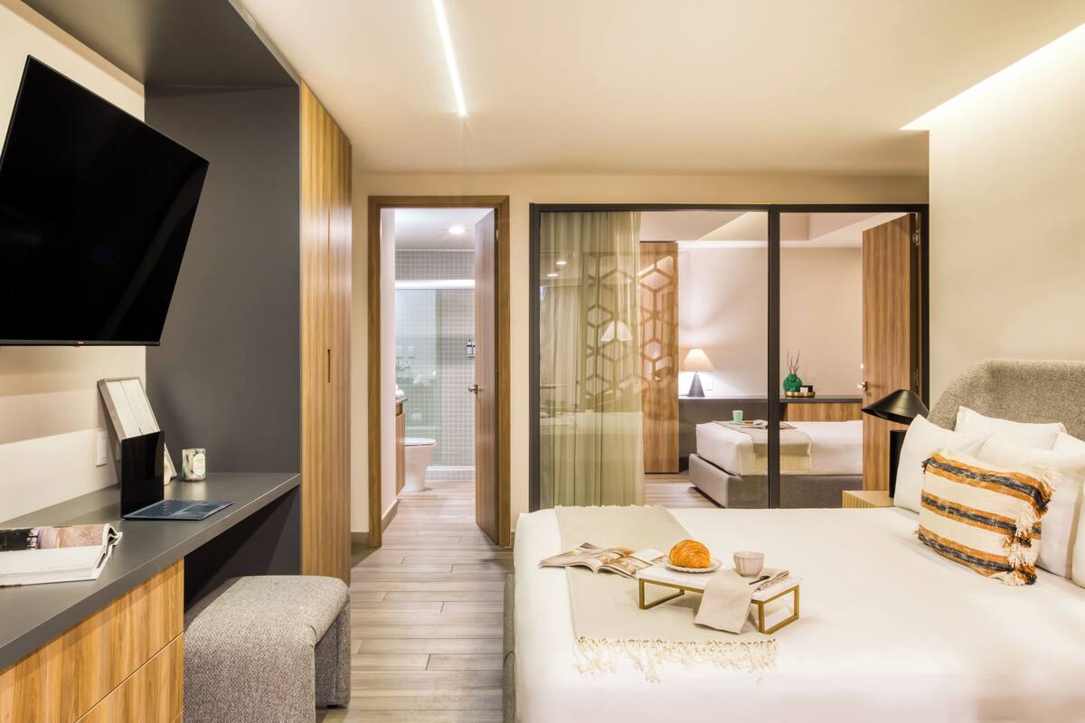 A modern hotel room with a flat-screen TV, a desk with a laptop, a bed with a bedside table, and a bathroom visible through an open door.
