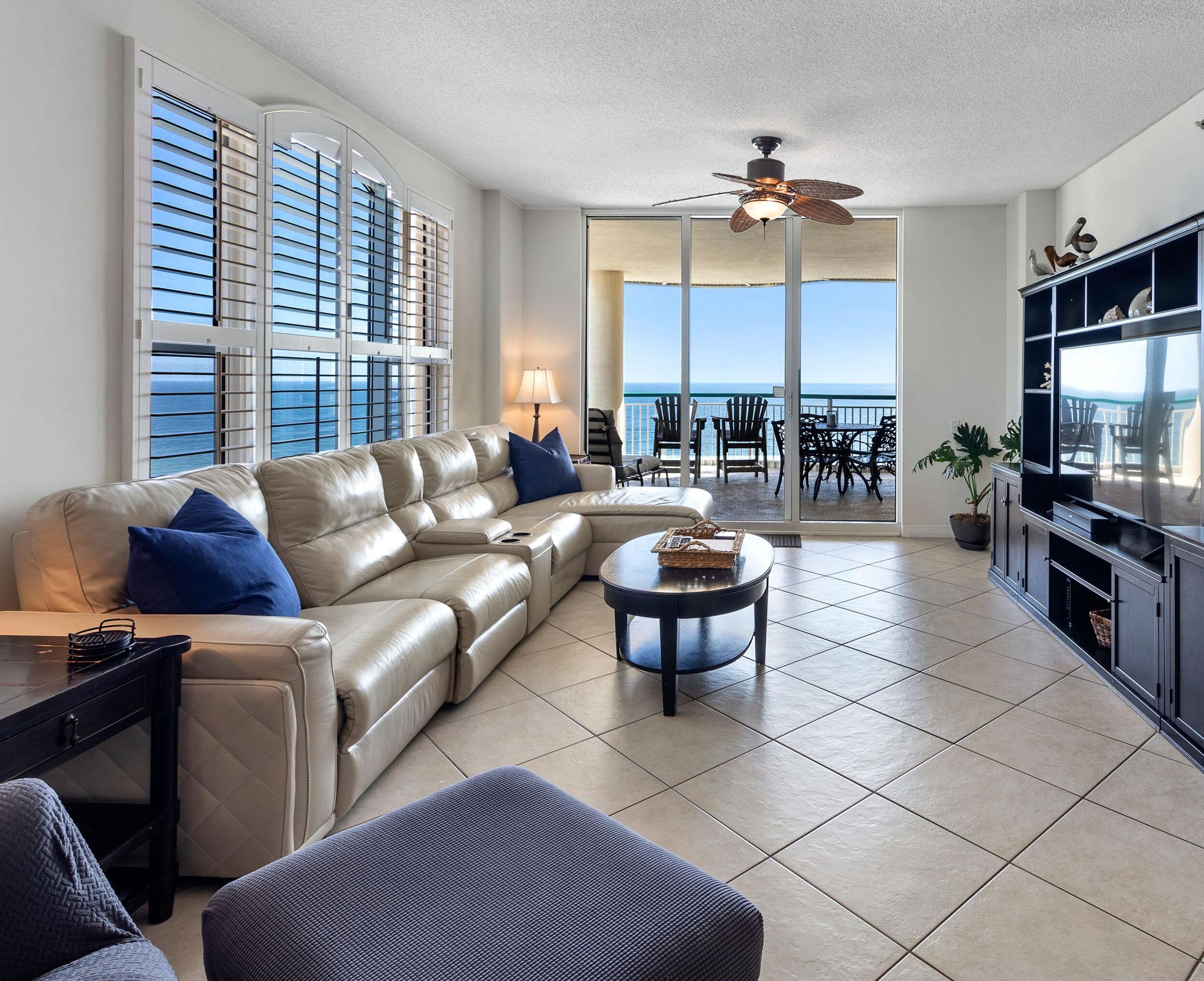 A spacious living room with a beige sectional sofa, a round coffee table, and a TV on a black stand. Large windows offer a view of the ocean.