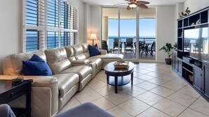 A spacious living room with a beige sectional sofa, a round coffee table, and a TV on a black stand. Large windows offer a view of the ocean. - Beach Colony East 10A (Pensacola)