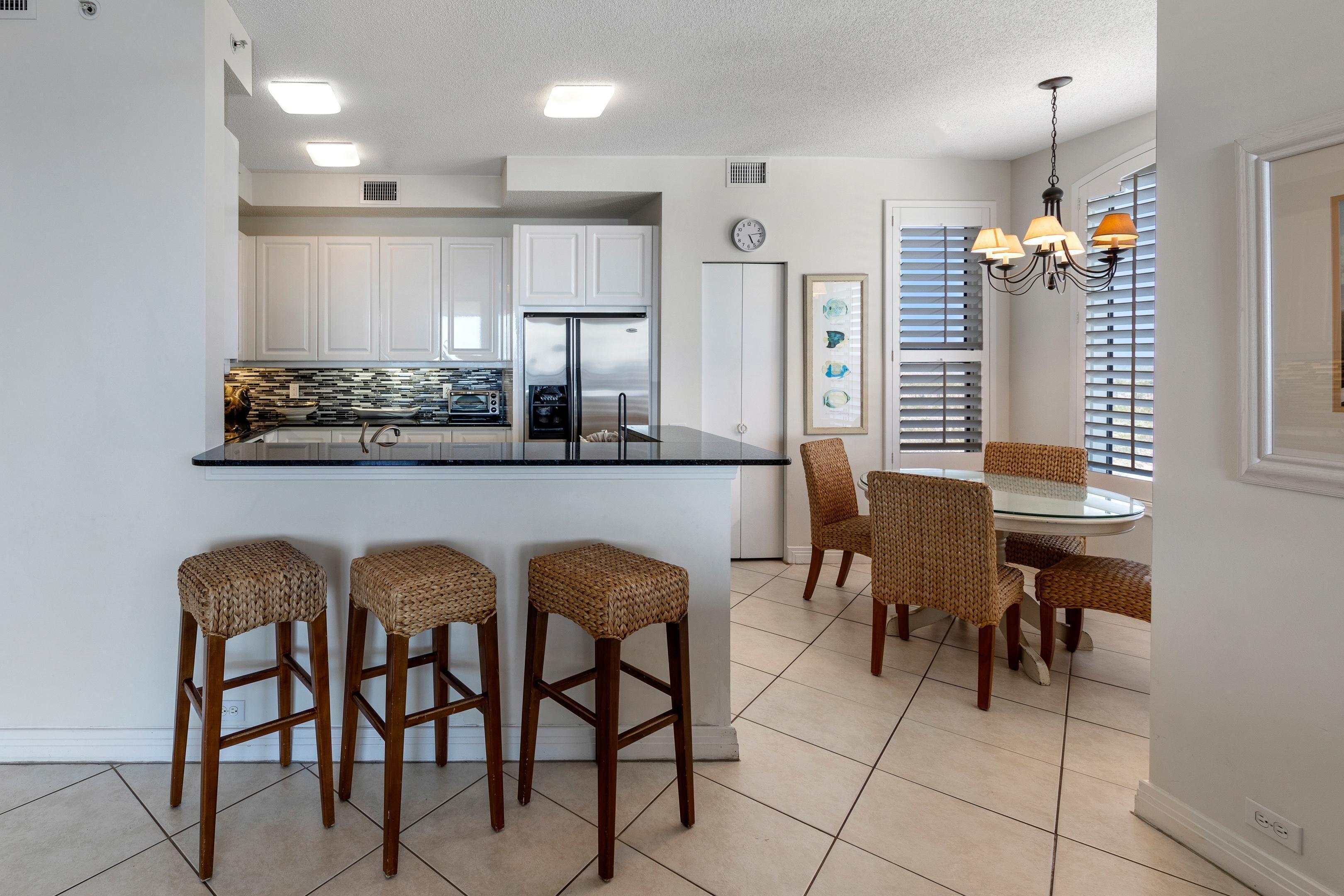 A modern kitchen with white cabinets, a stainless steel refrigerator, and a glass island with bar stools. There is a dining area with wicker chairs and a chandelier.