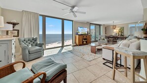 A spacious living room with a large window overlooking the beach, a ceiling fan, and a dining area with a view. - Perdido Towers West 908 (Pensacola)