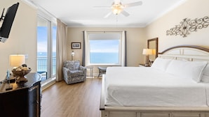 A hotel room with a large bed, a ceiling fan, a view of the ocean, a small table with a lamp, and a chair. - Indigo East 1405 (Pensacola)