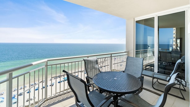 A balcony with a round table and chairs, overlooking a beach with umbrellas and a clear view of the ocean. - Indigo East 1405 (Pensacola)