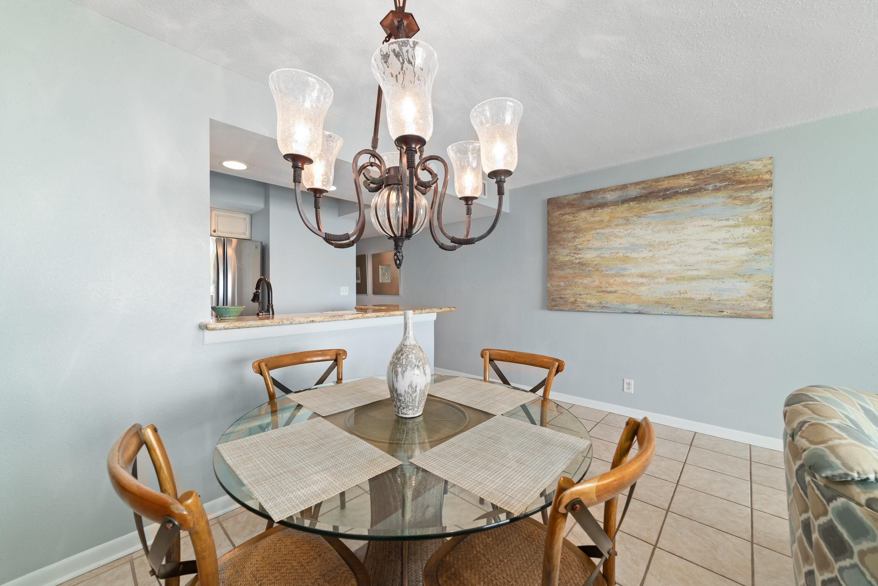 A dining area with a glass table, wooden chairs, a chandelier, a vase, and a painting on the wall.