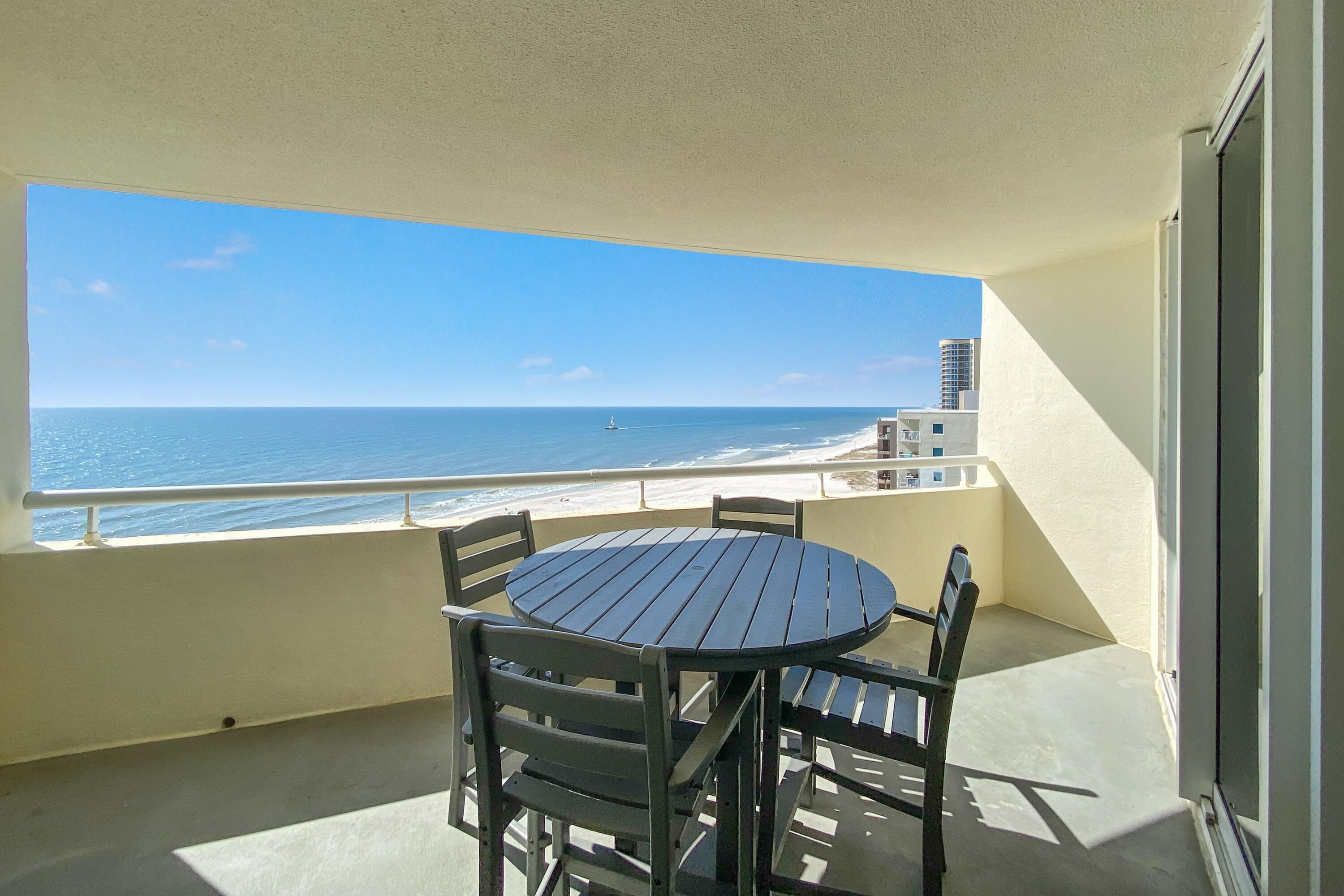 Ein Balkon mit rundem Tisch und Stühlen, der Blick auf einen Strand unter wolkenlosem Himmel gewährt.