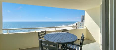Ein Balkon mit rundem Tisch und Stühlen, der Blick auf einen Strand unter wolkenlosem Himmel gewährt.