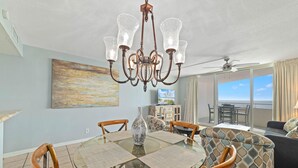 A dining area with a glass table, wooden chairs, a chandelier, and a painting on the wall. - Perdido Sun 904 (Pensacola)