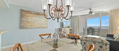 A dining area with a glass table, wooden chairs, a chandelier, and a painting on the wall.