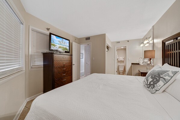 Un dormitorio con cama, televisión montada en una cómoda, escritorio con lámpara y baño visible a través de una puerta abierta.