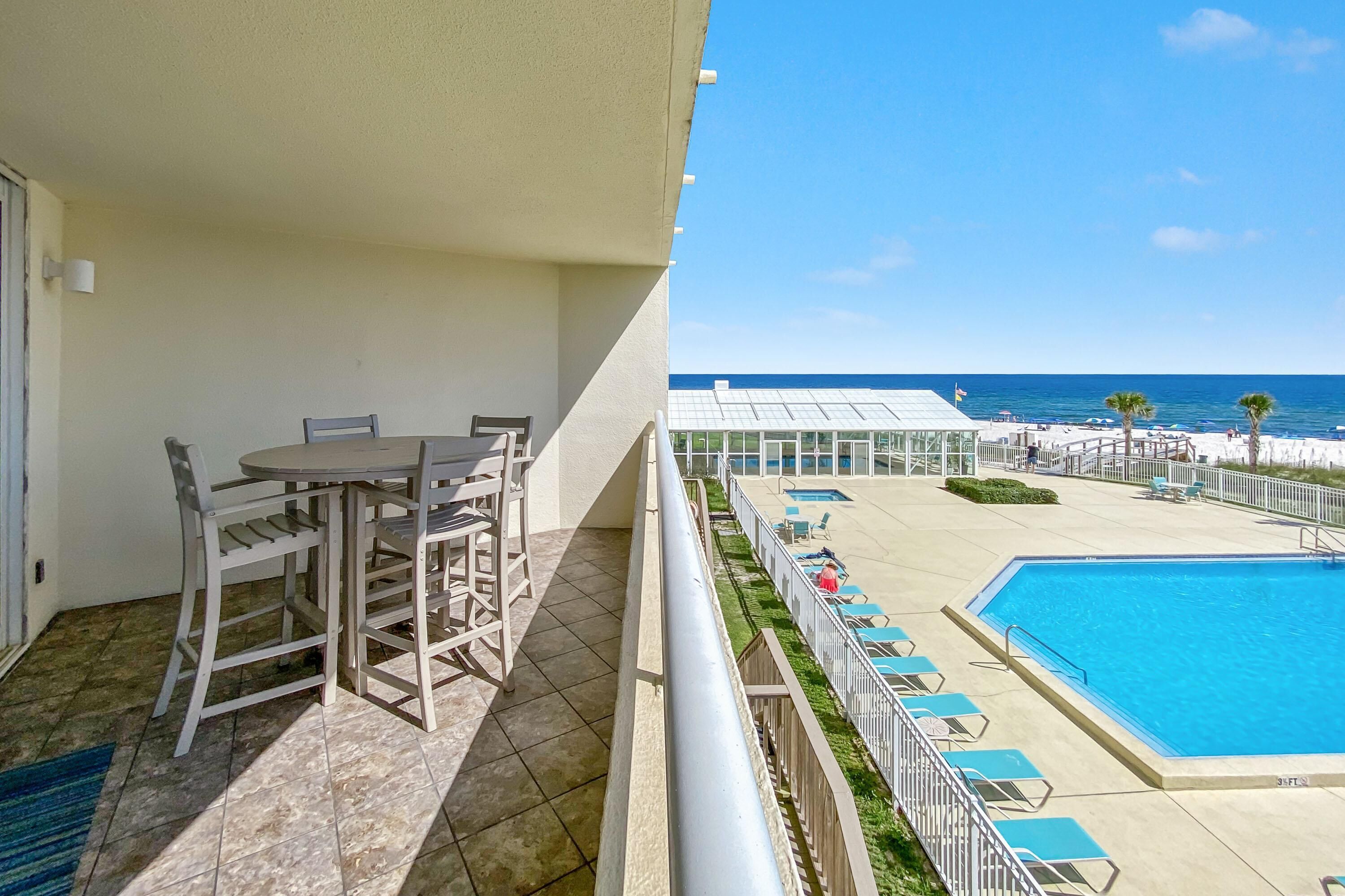 Un balcon avec une table et des chaises, une piscine avec des chaises longues, et une vue sur la plage et l’océan.