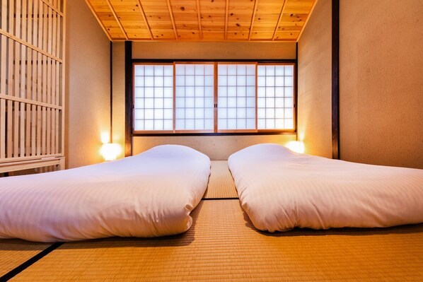 Una habitación tradicional japonesa con suelo de tatami, dos camas con ropa blanca, una ventana con pantalla shoji y vigas de madera en el techo.