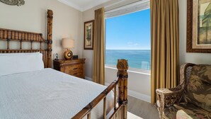 Een hotelkamer met een groot bed, een houten hoofdeinde, een nachtkastje met een lamp, een stoel en uitzicht op de oceaan.
