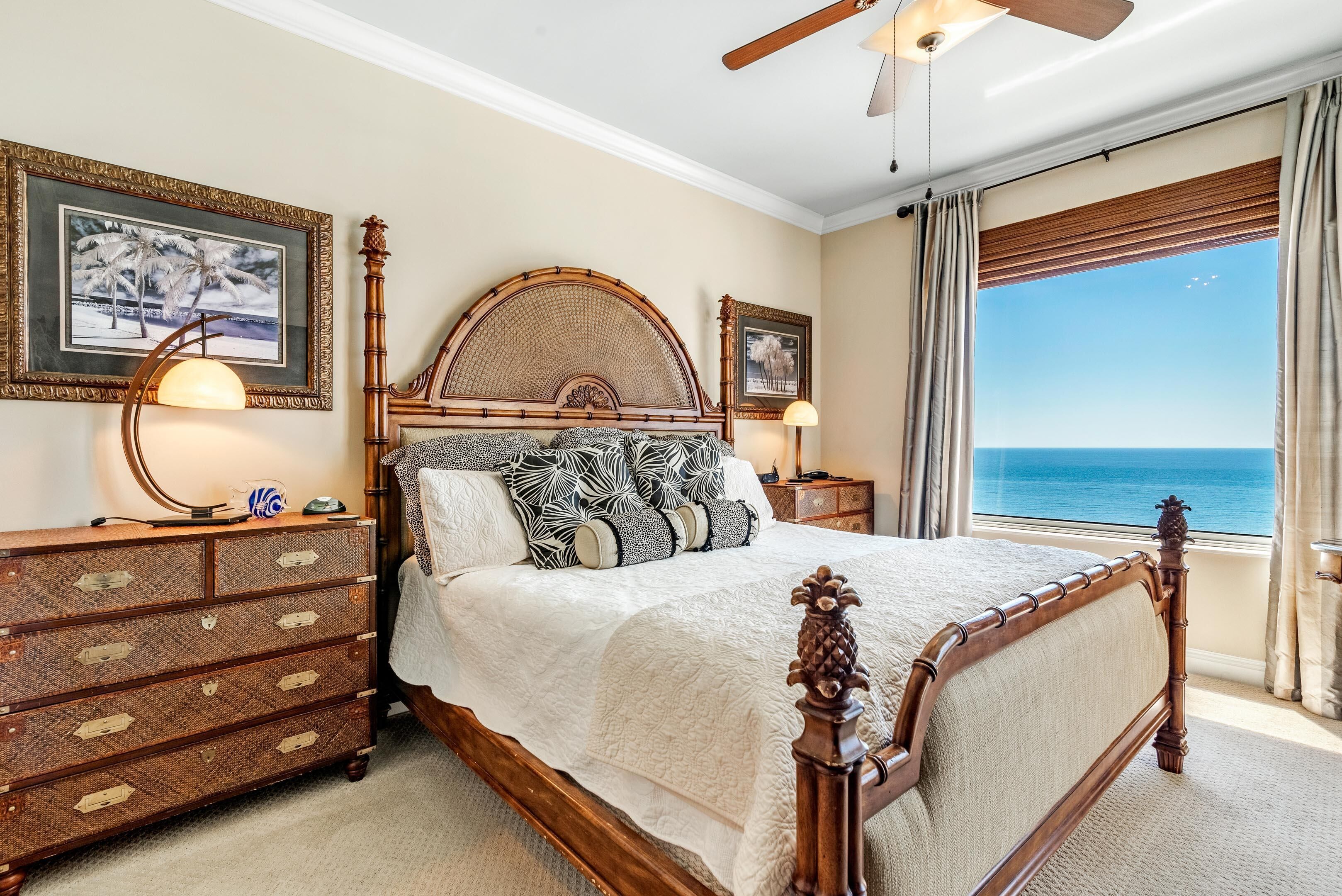 A bedroom with a large bed, wooden headboard, a dresser, and a view of the ocean through a window.