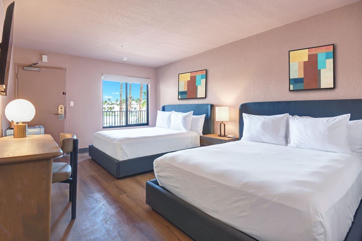 A hotel room with two beds, a desk, a chair, a window with a view of palm trees, and abstract wall art.