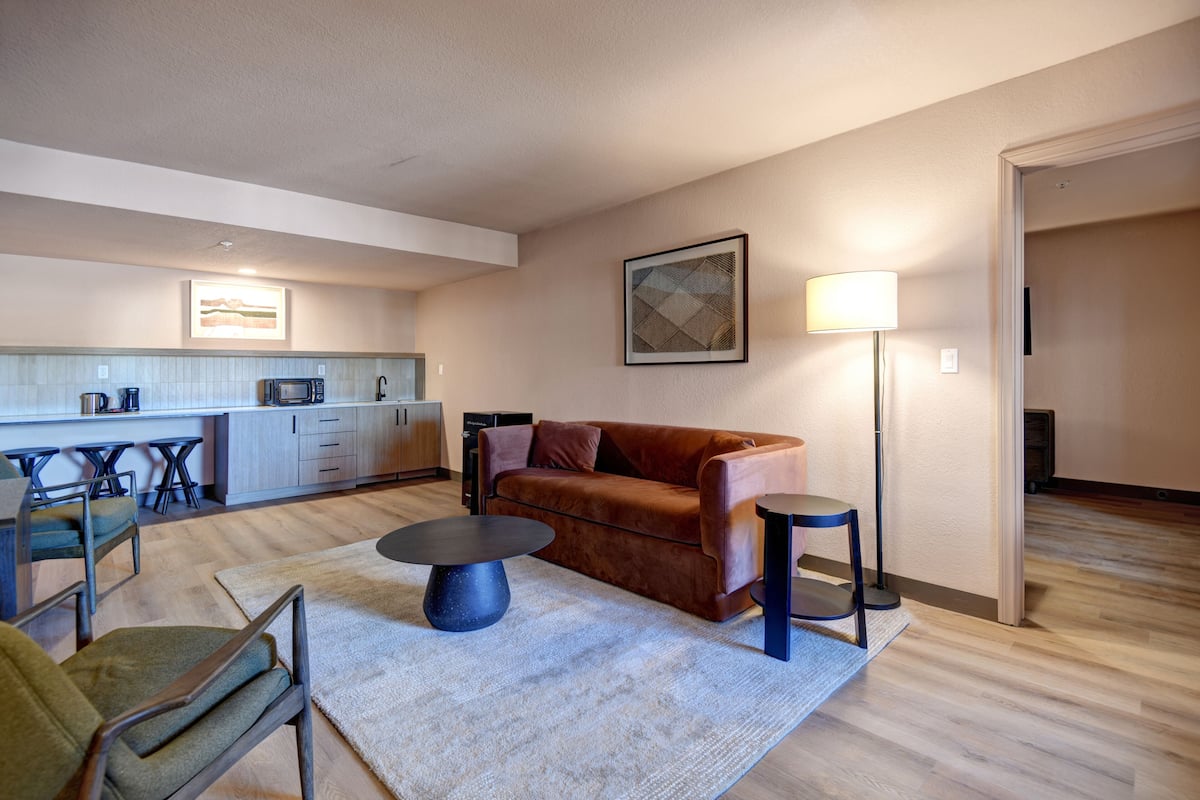 A modern living room with a brown sofa, a round coffee table, and a floor lamp. A kitchen with a microwave and bar stools is visible in the background.