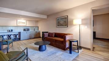 A modern living room with a brown sofa, a round coffee table, and a floor lamp. A kitchen with a microwave and bar stools is visible in the background.