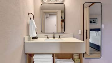 Une salle de bain avec un lavabo blanc, trois lampes au-dessus et un miroir. Des serviettes sont posées sur une étagère située sous le lavabo.