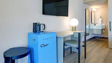 A compact hotel room with a kitchenette, a flat-screen TV mounted on the wall, a small round table, a blue refrigerator, a chair, and a mirror above the sink.