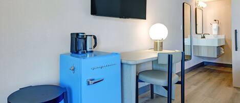 A compact hotel room with a kitchenette, a flat-screen TV mounted on the wall, a small round table, a blue refrigerator, a chair, and a mirror above the sink.