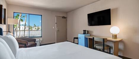 A hotel room with a large bed, a desk with a lamp, a television, and a window with a view of palm trees.