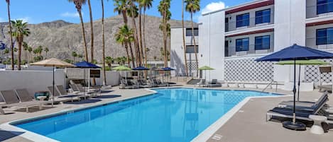 A swimming pool with lounge chairs and umbrellas, surrounded by palm trees and a building with balconies.