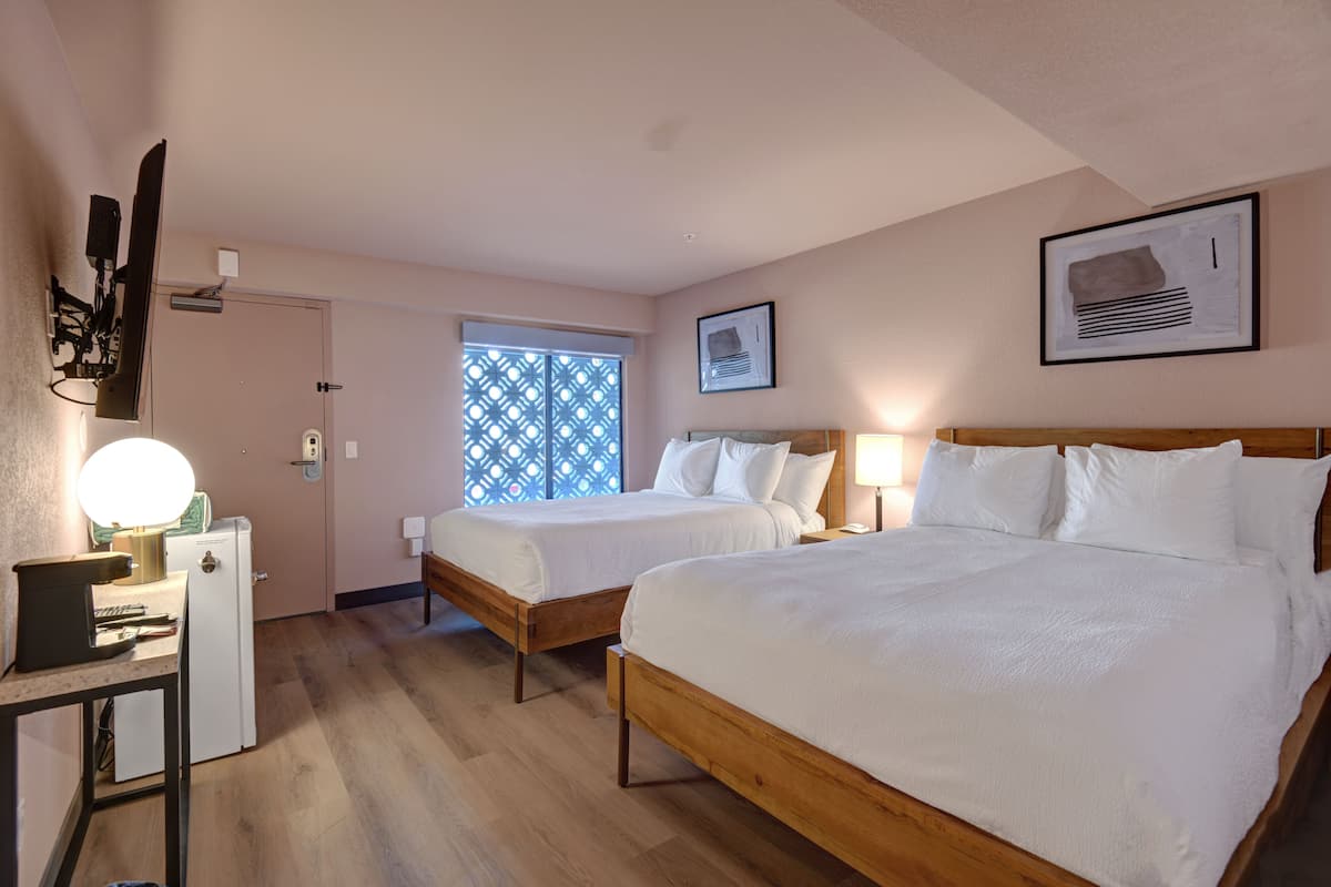 A hotel room with two single beds, a desk with a lamp, a small fridge, and a window with a lattice design.