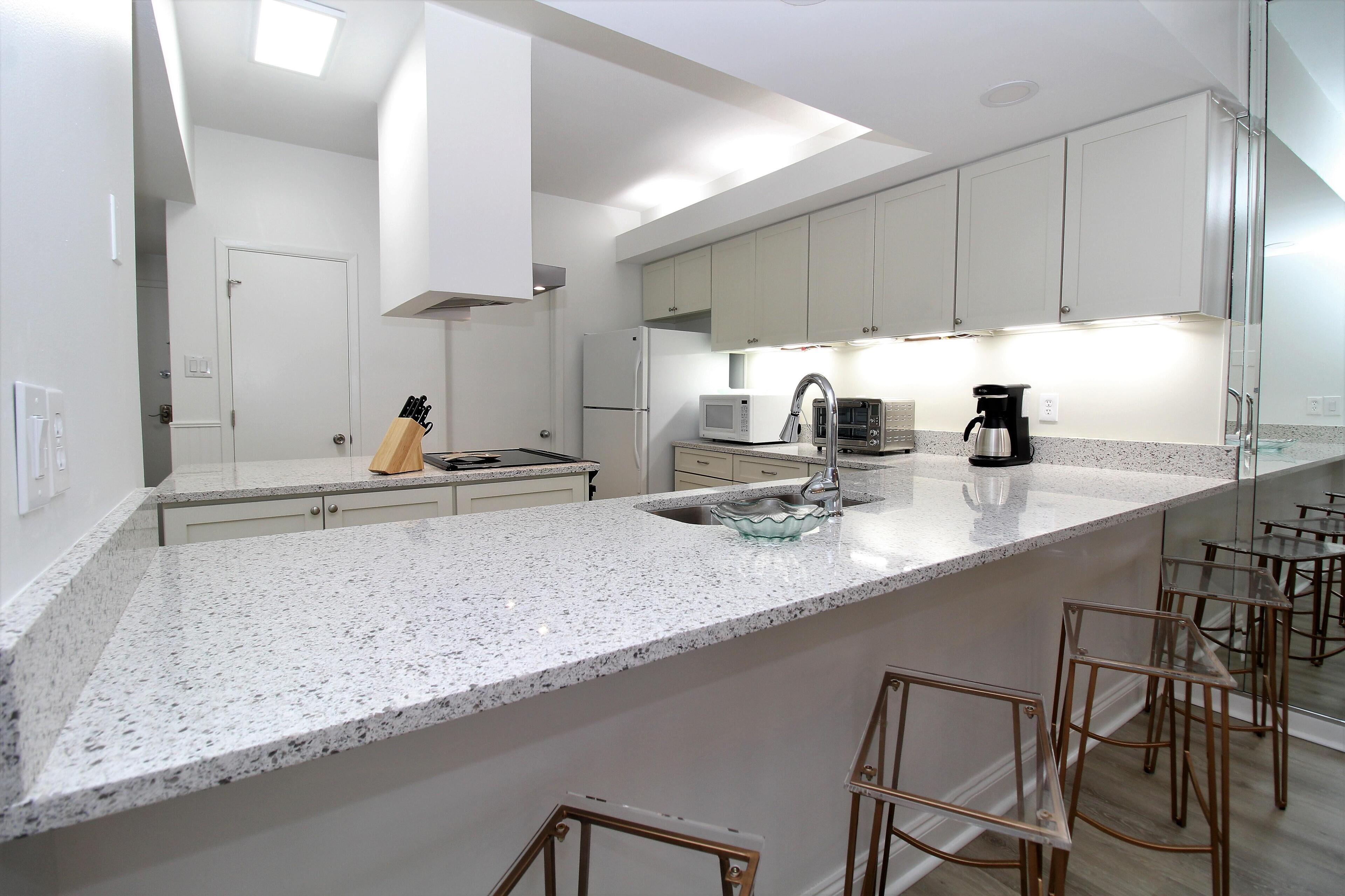 Eine moderne Küche mit weißen Schränken, einer Granit Arbeitsplatte, einer Edelstahl-Spüle, einer Mikrowelle, einer Kaffeemaschine und Barhocker.