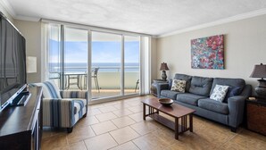 A living room with a large TV, a sofa, a coffee table, and a balcony with a view of the ocean.
